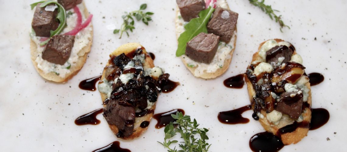 Four pieces of baguette with steak cubes, blue cheese, greens, balsamic reduction